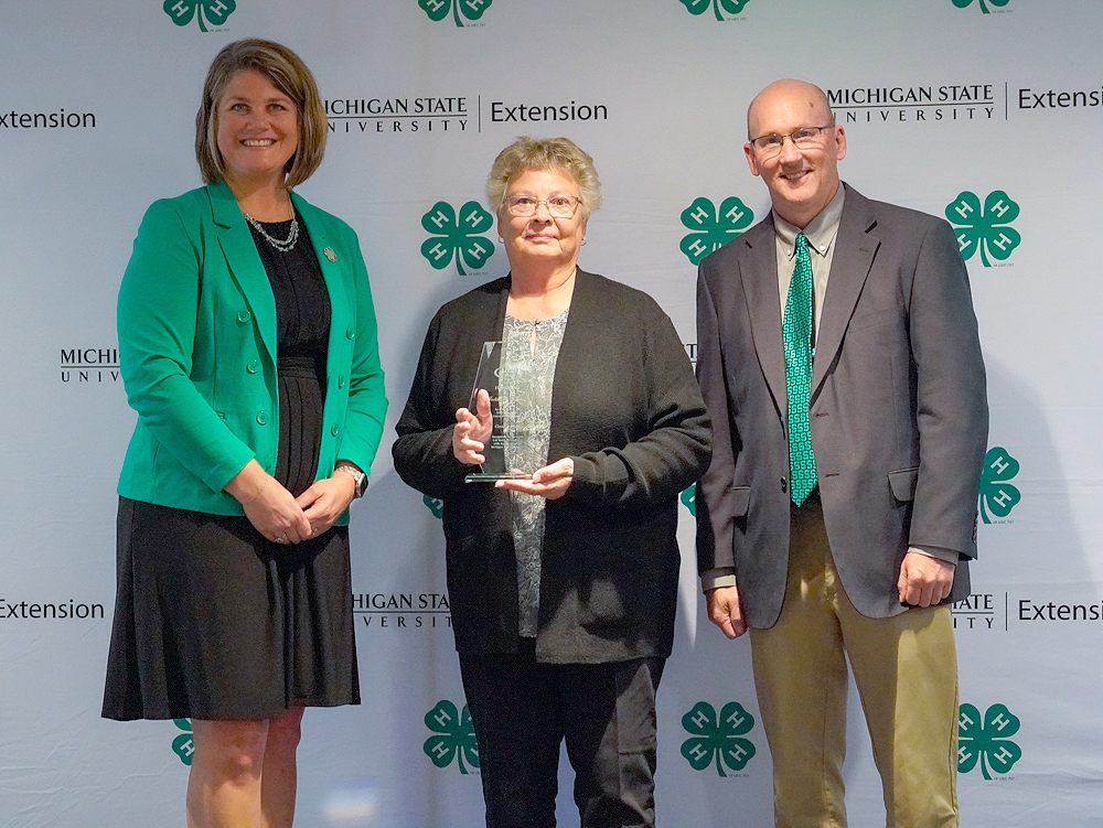 Three individuals at a backdrop. The center person holds an award.